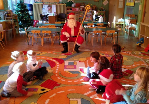 Mikołaj siedzi na krześle na środku sali. Dzieci siedzą w kole na dywanie, rozmawiają z Mikołajem, czekają na prezent.