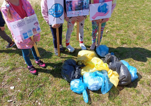 Dzieci trzymają w rękach tabliczki z plakatami związanymi z Dniem Ziemi i akcją "Sprzątamy dla Polski". Obok leżą worki w różnych kolorach ze śmieciami.