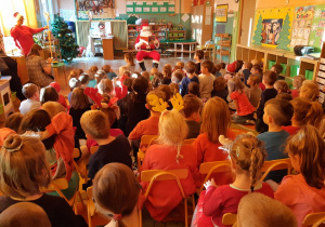 Mikołaj siedzi na środku sali i czyta książkę. Dzieci siedzą na dywanie, obserwują i słuchają. Mikołajka stoi przy oknie.
