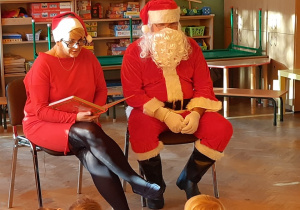 Mikołaj i Mikołajka siedzą na krzesłach. Mikołajka czyta książkę i spogląda w stronę dzieci.