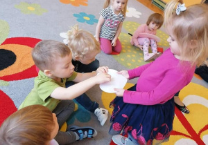 Dzieci siedzą na dywanie. Dziewczynka trzyma w rączkach serce z imionami. Dzieci nakłuwają serce i losują imię najlepszego kolegi lub koleżanki.