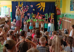Na scenie kobieta i dzieci, którzy w rękach trzymają litery tworzące napis wakacje.