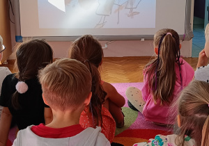 Dzieci siedzą na dywanie i oglądają na tablicy multimedialnej film o małej Vashti.