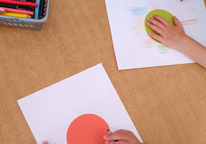 Dzieci siedzą przy stoliku. Z kolorowych kropek przyklejonych na kartce powstają kreatywne dzieła wykonane przez dzieci.