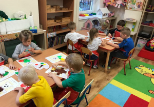 Dzieci siedzą przy dwóch stolikach, malują misia farbami przy pomocy gąbek.