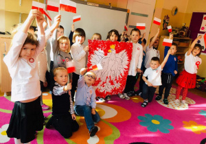 Dzieci stoją w sali, prezentują polskie flagi i godło.