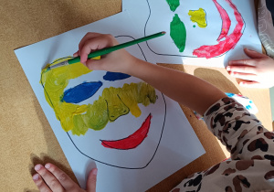 Dzieci przy stoliku malują farbami maski teatralne z wykorzystaniem pędzelka.