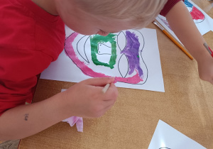 Chłopiec przy stoliku maluje farbami maskę teatralną z wykorzystaniem pędzelka.