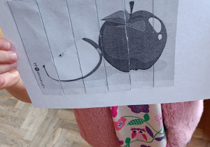 Dziewczynka trzyma w rączkach wykonane zadanie tygodnia - obrazek pocięty i przyklejony na kartkę przedstawiający jabłka.