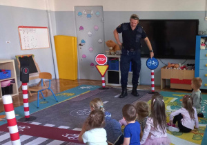 Strażnik Miejski stoi na imitacji ulicy i opowiada dzieciom o znakach drogowych.