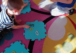 Dzieci siedzą na dywanie i na piankowym puzzlach układają literki z darów zabawy Froebla.