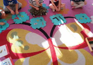 Dzieci siedzą na dywanie i prezentują literki ułożone na piankowych puzzlach z darów zabawy Froebla.