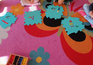 Dzieci siedzą na dywanie i prezentują literki ułożone na piankowych puzzlach z darów zabawy Froebla.