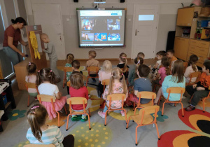 Dzieci siedzą na krzesełkach przed tablicą multimedialną.