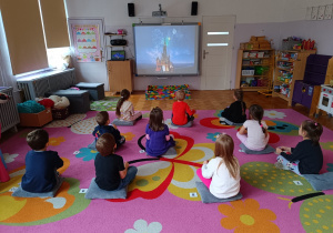 Dzieci siedzą na dywanie przed monitorem multimedialnym, na którym rozpoczyna sie bajka.