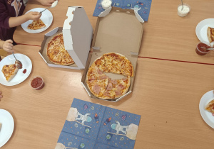 Dzieci z grupy czerwonej przy stoliku jedzą pizzę.