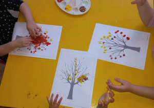 Dzieci malują palcami jesienne liście na drzewach.