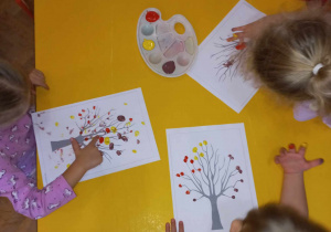 Dzieci malują palcami jesienne liście na drzewach.