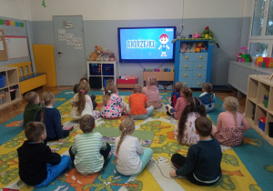 Dzieci oglądają na monitorze multimedialnym film edukacyjny o andrzejkach.