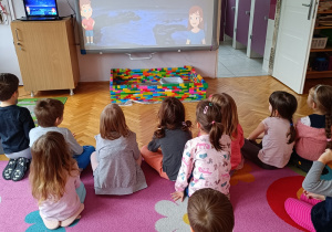 Dzieci siedzą na dywanie i oglądają film edukacyjny o andrzejkach na monitorze multimedialnym.