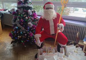 Mikołaj siedzi na krześle obok choinki, przed nim prezenty dla dzieci.