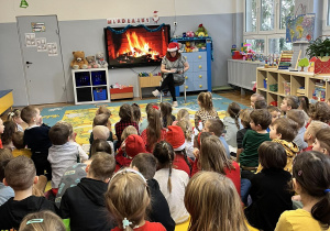 Dzieci siedzą na dywanie. Nauczycielka w czapce Mikołaja czyta przedszkolakom wiersze związane z tematyką świąteczną.