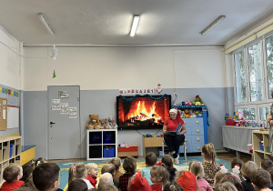 Dzieci siedzą na dywanie. Nauczycielka w czapce Mikołaja czyta przedszkolakom wiersze związane z tematyką świąteczną.