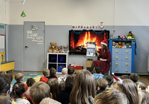 Dzieci siedzą na dywanie. Nauczycielka w czapce Mikołaja czyta przedszkolakom opowiadania związane z tematyką świąteczną.