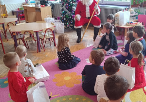 Dzieci siedzą na dywanie przed Mikołajem i śpiewają mu piosenkę.