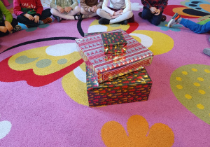 Dzieci siedzą na dywanie wokół zapakowanych prezentów.