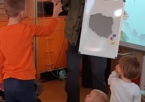 Pisarz stoi przed dziećmi, prezentuje mapę. Chłopiec wskazuje miejsce na mapie.