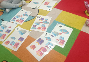 Dzieci siedzą na dywanie, w środku ilustracje przedstawiające różną liczbę prezentów oraz odpowiadające im cyfry.