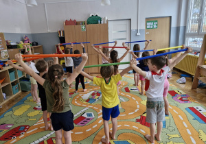 Dzieci stoją z laskami gimnastycznymi uniesionymi do góry.