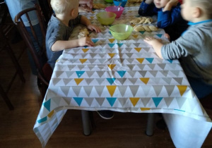 Dzieci siedzą przy stole, Pani pomaga im wyrabiać ciasto.