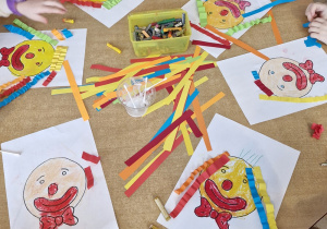 Dzieci przy stoliku tworzą fryzury dla karnawałowych klaunów - zaginają paski kolorowego papieru i przyklejają je na głowie klauna.