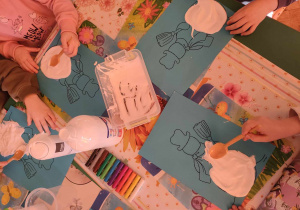 Dzieci malują białą farbą bałwana na niebieskiej kartce.