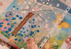 Chłopiec palcem maluje białą i niebieską farbą drzewo.