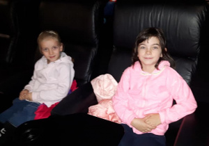 Dziewczynki siedzą na miejscach w sali kinowej i uśmiechają się.