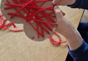 Dziecko przewleka czerwoną włóczkę przez nacięcia zrobione w tekturowym serduszku.