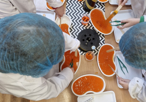 Dzieci pracują przy stoliku - przy pomocy drewnianych patyczków, plastikowych noży i pęsety próbują wydostać z galaretki sensorycznej monetę.