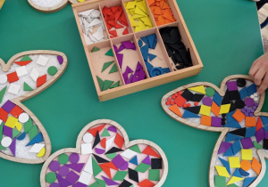 Dzieci układają kolorowe dary Froebla na drewnianych konturach kwiatka, motyla i listka.