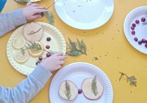 Dzieci tworzą swoje kompozycje z owoców i gałązek na papierowych talerzach.