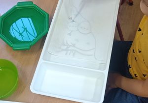 Dziecko przy stoliku z wykorzystaniem pipety i wody zakrapia obrazek znajdujący się w plastikowym pojemniku.