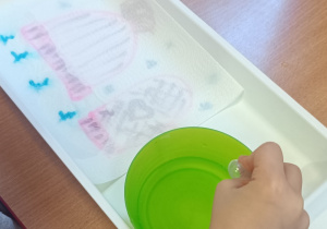 Dzieci przy stoliku z wykorzystaniem pipety i wody zakrapiają obrazki znajdujące się w plastikowych pojemnikach.