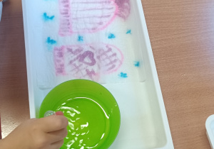 Dziecko przy stoliku z wykorzystaniem pipety i wody zakrapia obrazek znajdujący się w plastikowym pojemniku.