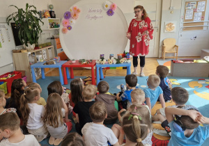 Nauczycielka opowiada dzieciom siedzącym na dywanie o kompozycjach z różnych roślin.