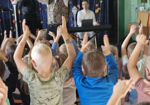 Dzieci na ławeczkach, podnosząc wysoko ręce, klaszczą w rytm muzyki.