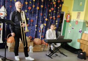 Na scenie mężczyźni grają na instrumentach: na puzonie i na pianinie.