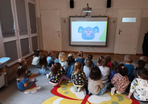 Dzieci siedzą na dywanie i oglądają na tablicy multimedialnej film edukacyjny o prawach dziecka.