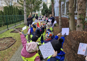 Dzieci ustawione w parach przed przedszkolem w kamizelkach odblaskowych podnoszą do góry przygotowane przez siebie kartki świąteczne.